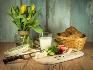 Tisch mit Blumenstrauß, Milch, einen Korb Brötchen und einem Brotbrett mit Käse, Gurke, Tomaten, Petersilie und Salz vor einem Glas Milch
