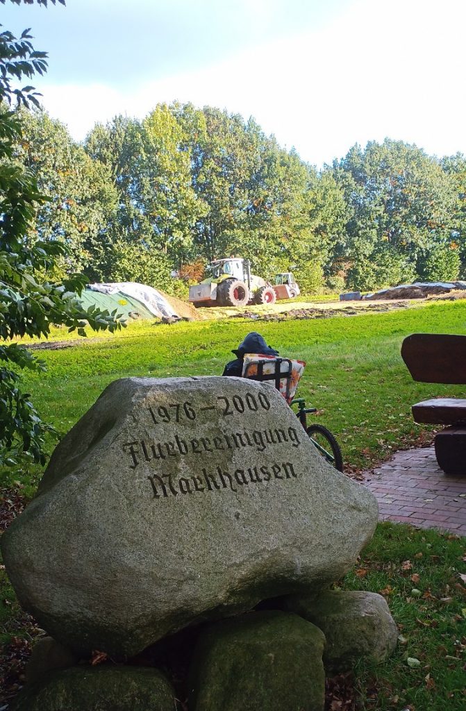 Im Hintergrund ein Trecker an einem Silo, dann Wiese, hinter einem großen Gedenkstrein von der Flurbereinigung in Markhausen sieht man Nick auf dem Sacco